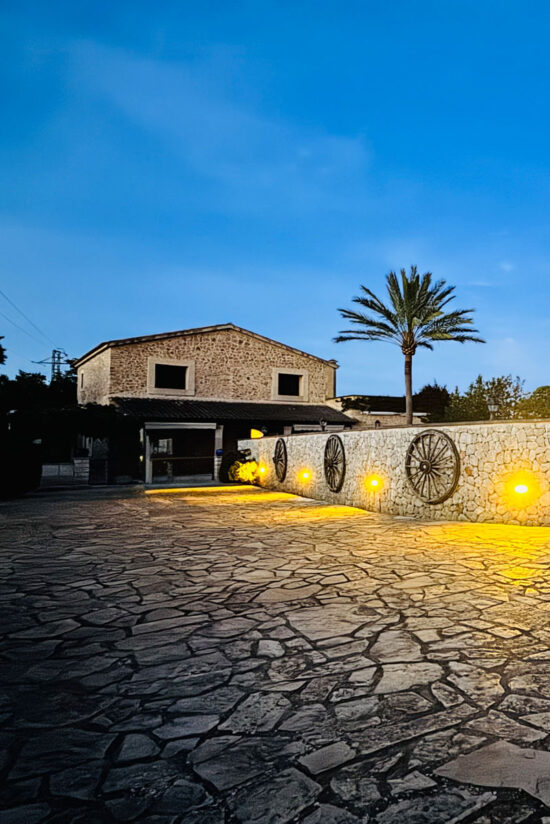 Patio empedrado de Son Fullaneta al anochecer, con la casa de piedra, una palmera y antiguas ruedas de carro iluminadas sobre el muro, Llucmajor